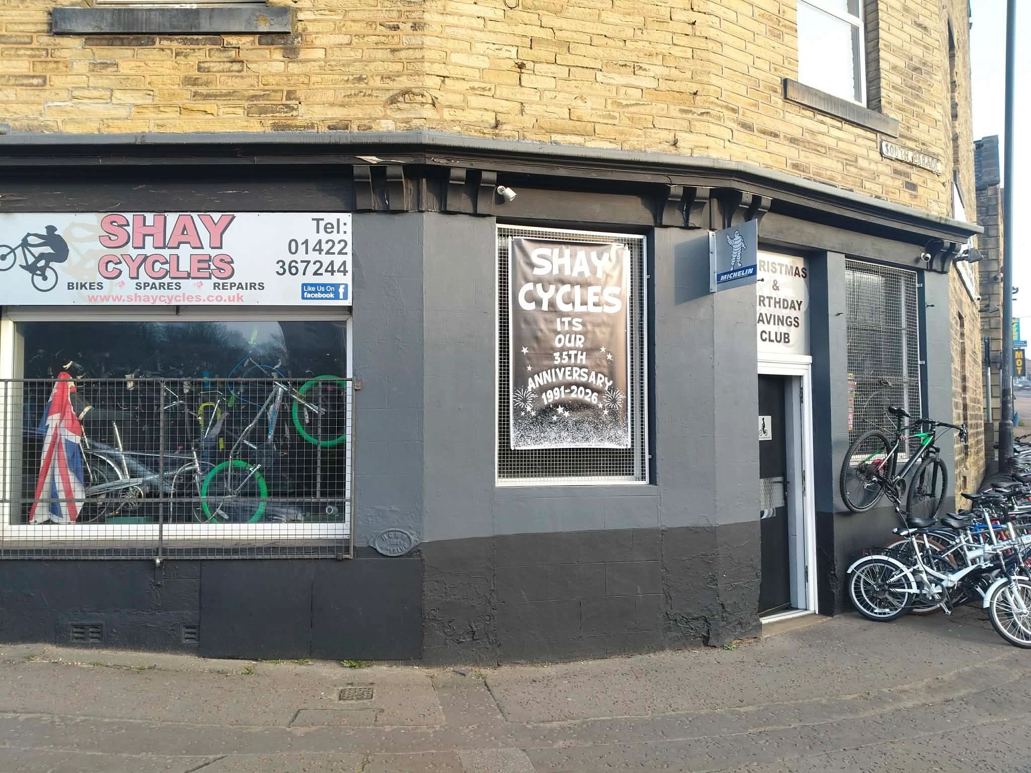 The full Shay Cycles shopfront on South Parade, with its black-painted stone frontage, red and white sign, mesh windows, anniversary poster and bikes outside